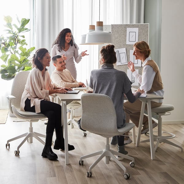 Five discuss at a modern office desk, one pointing. Bright room, large window.