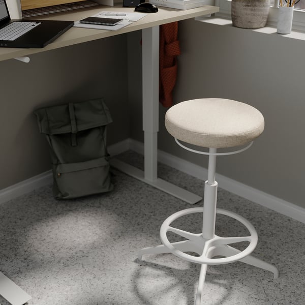 Home office setup: white adjustable stool, beige cushioned seat, grey desk with laptop, mouse, and backpack.
