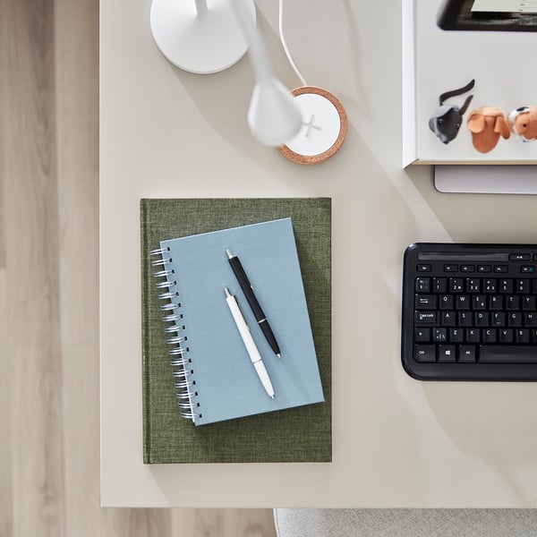 A minimalist desk setup with a green notebook, pens, keyboard, and lamp, emphasizing the TROTTEN desktops size and design.