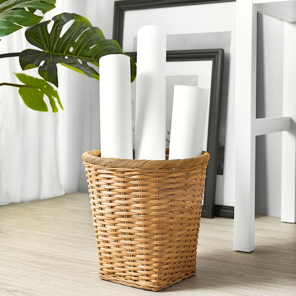 Wicker basket TRUMMIS holding white items, set on wood floor with plant and frames.