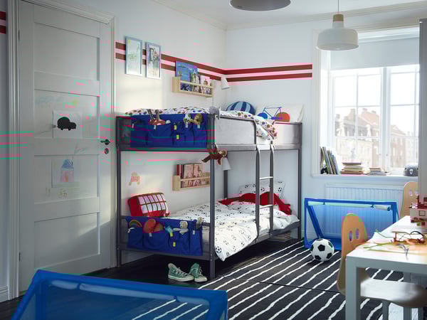 Kids room with blue-striped wall, bunk bed with sports theme, black and white striped rug, blue storage, wooden chair, and books by window.