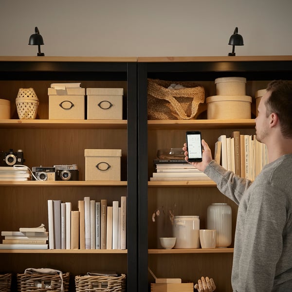 A person adjusts the person's phone, illuminating a wooden bookshelf with integrated, adjustable cabinet lighting, highlighting items stored inside.