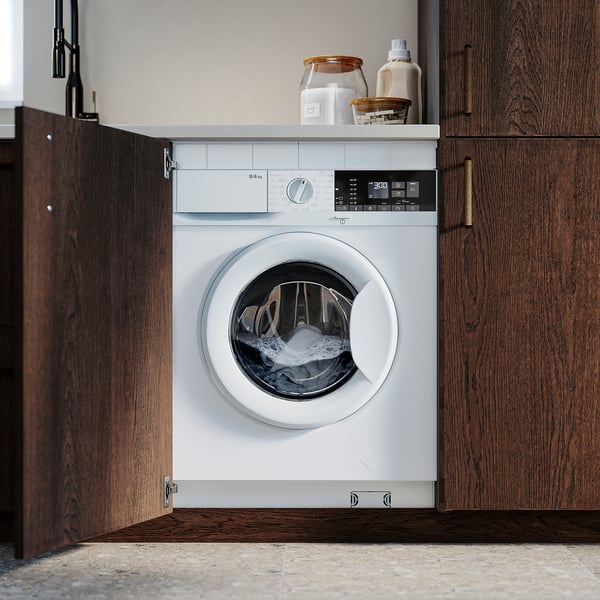 White front-loading washer/dryer combo installed in wooden cabinet space, with control panels and laundry detergent on top.