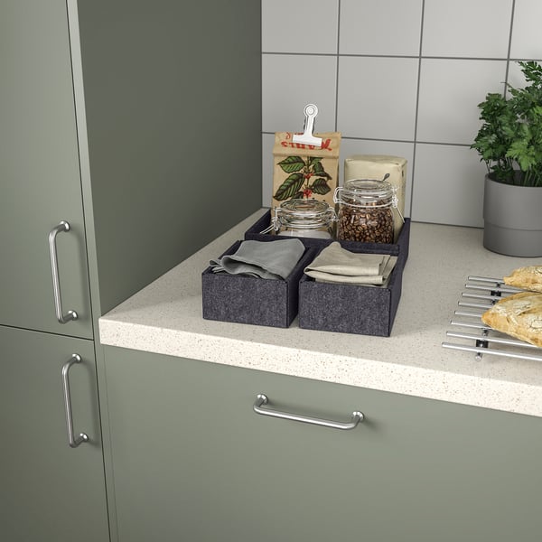 Kitchen counter with green cabinets and speckled countertop. Two grey boxes with cloth napkins, jars, and a paper bag with coffee beans and clips.