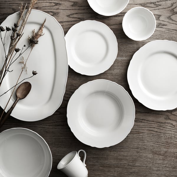 White rustic porcelain dinnerware set includes plates, bowls, and mugs with ruffled edges on a wooden surface, dried flowers added for decoration.
