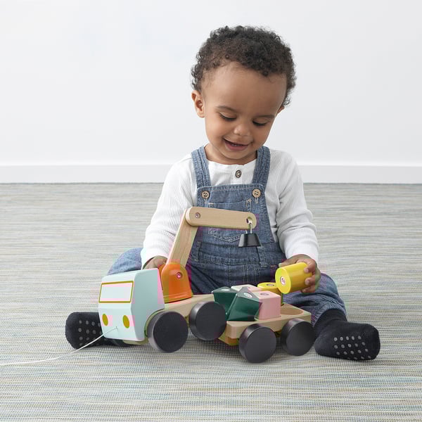 Child playing with wooden pull toy truck and crane on rug. Truck has large wheels and magnetic crane.