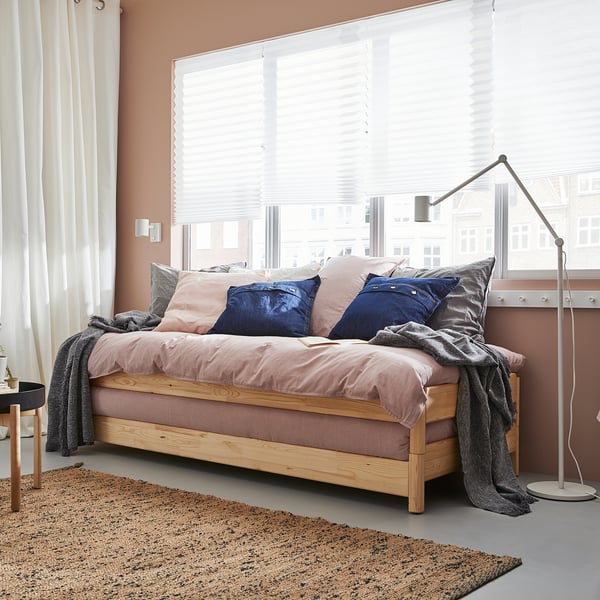 Bedroom with UTÅKER bed, light pink linen, blue pillows, wooden frame, floor lamp, rug, and large window with blinds.