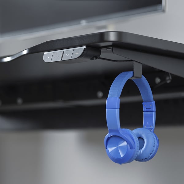A black gaming desk with a control panel for adjusting height. Blue headphones hang from a hook on the side.
