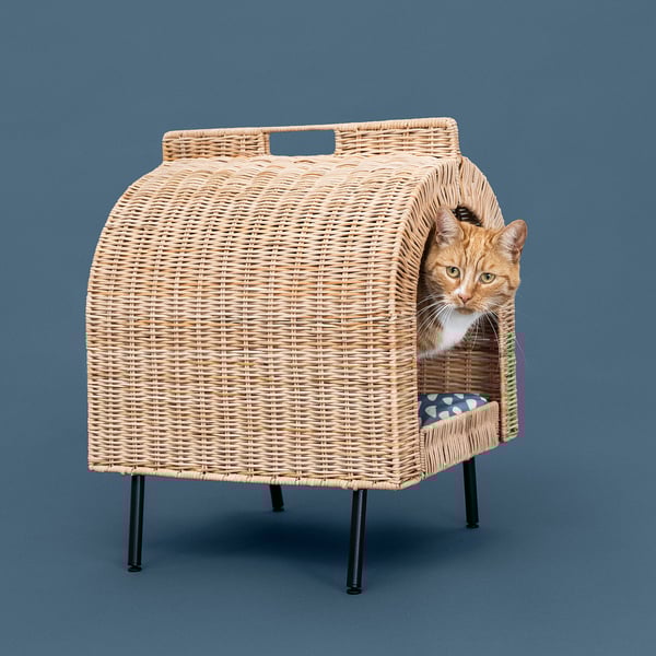 A wicker cat house with a cushion inside, elevated on four legs. A cat peeks out from the entrance.