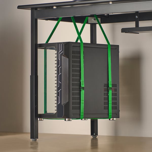 Black gaming desk with green straps holding computer tower under shelf, featuring cable management and height adjustment.