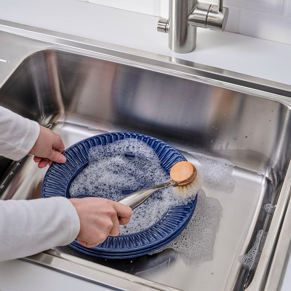 Person washing blue dish in sink using dish brush and soap.