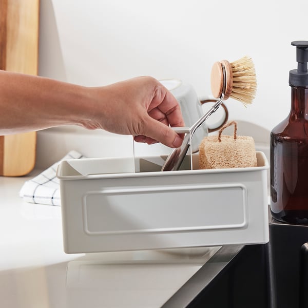 Hand placing a scrub brush into a white sink organiser with compartments and handle.