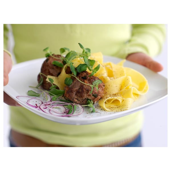 Person holding white square plate with soft edges, featuring grilled meat, yellow sauce, pasta, and purple onions garnished with green herbs.