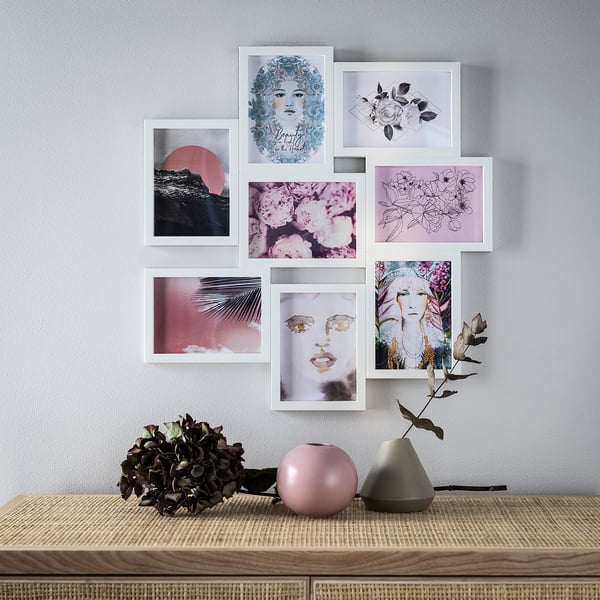 This image a wall display of seven VÄXBO frames in white, housing various artistic pictures. Below, a wooden table holds two vases and dried plants.