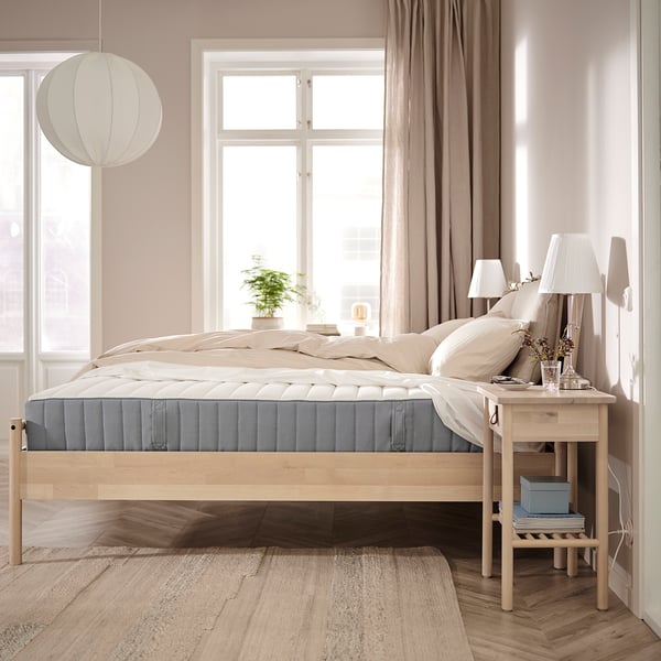 Bedroom with wooden floor, beige walls, large window & VALEVÅG mattress. Beside table has a lamp & plant, and a pendant lamp hangs nearby.