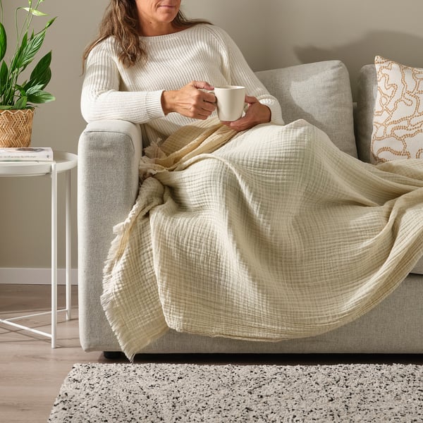 Person relaxes on couch with VALLKRASSING throw blanket, sipping a drink. Soft cotton muslin with crinkled effect and raw edges.