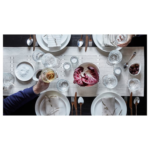 A hand raises a wine glass above a neatly set table with the VÅRARV table runner, white plates, glasses, cutlery, and decorations.