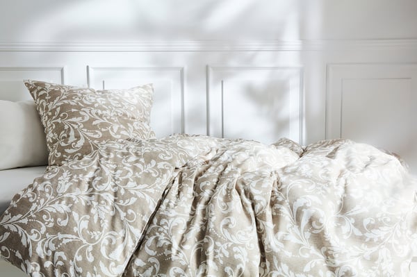 Bed with cream-colored VÅRBRÄCKA quilt and pillow, featuring intricate floral patterns, against a white headboard.