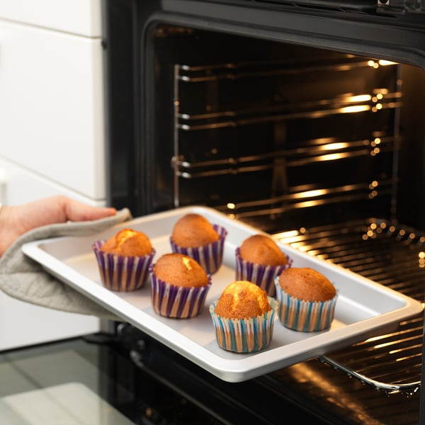 Person removing muffin tray from oven. Muffin tray has colourful striped cups with baked muffins. Tray is placed on oven rack.