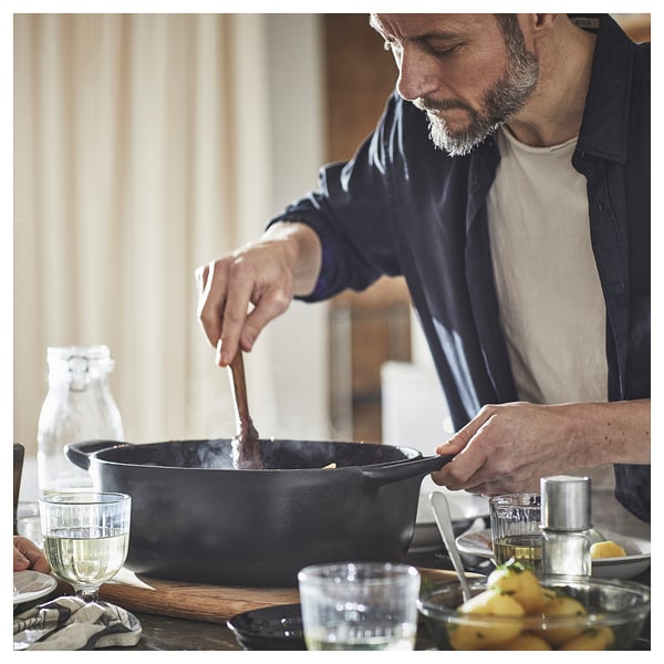 Person cooks in a black VARDAGEN cast iron pan on a wooden table with glasses and utensils.