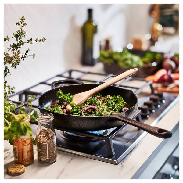 Black cast iron frying pan with wooden handle on stove, surrounded by spices and vegetables.