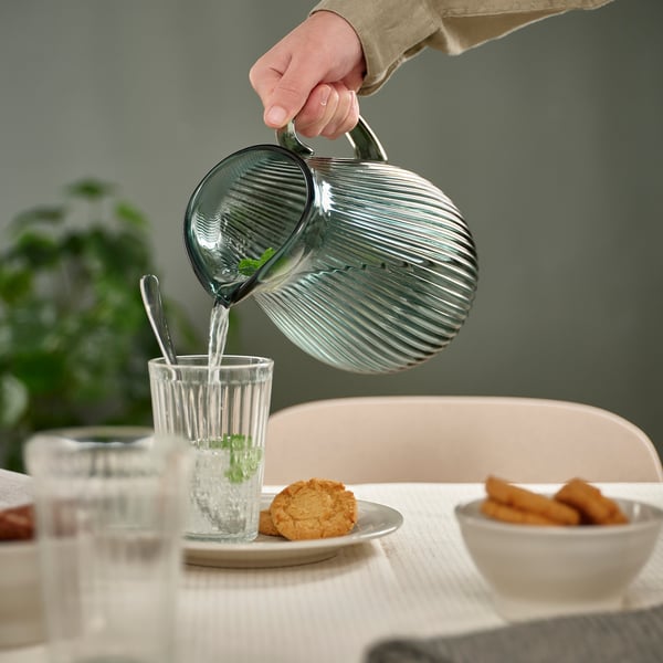 Hand pours water from a ribbed glass pitcher into a tall glass beside plates with cookies and fried snacks on a table.