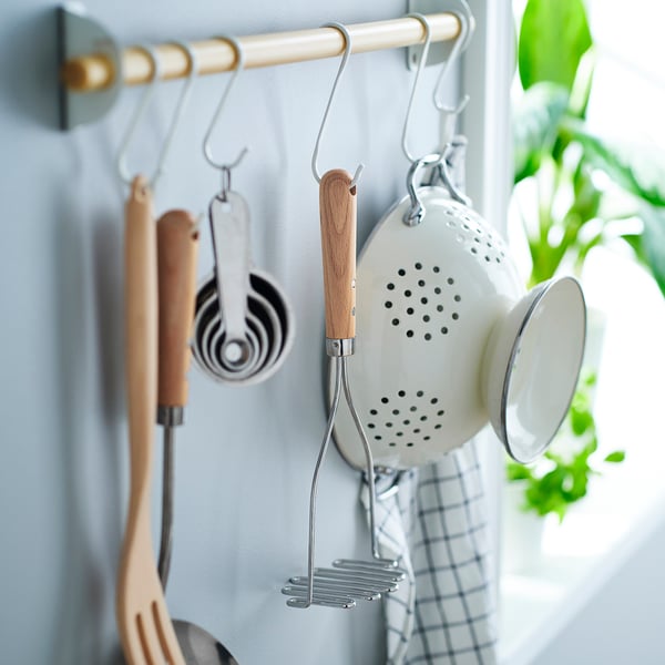 Kitchen utensils hang on a rack. Includes masher, strainer, measuring cups, and spoons. Wooden handles for grip.