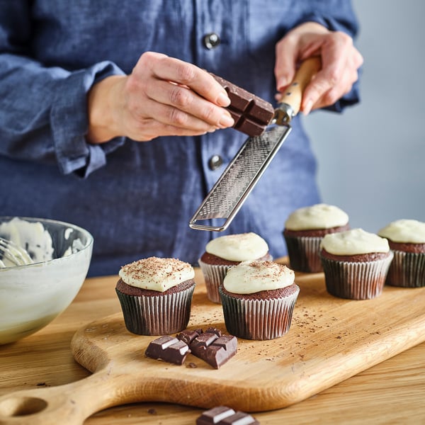 Person grates chocolate over frosted cupcakes on a wooden board for decoration. A bowl of icing is nearby.
