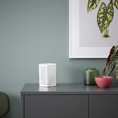 Small white VÅRSYREN lantern, steel, mesh design, sits on dark cabinet next to green and pink pots, framed plant poster behind.