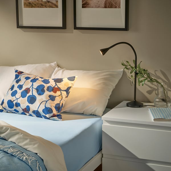 A bed with a light blue VÅRVIAL cotton pillow, white pillows, and a simple black lamp on a small white bedside table.