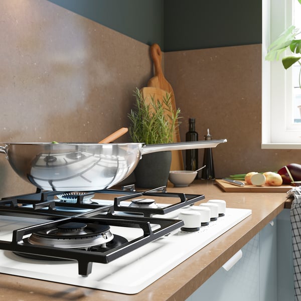 A modern kitchen with a white gas stove, featuring a wok on one burner. Nearby are herbs, bottles, and fruits on the counter.