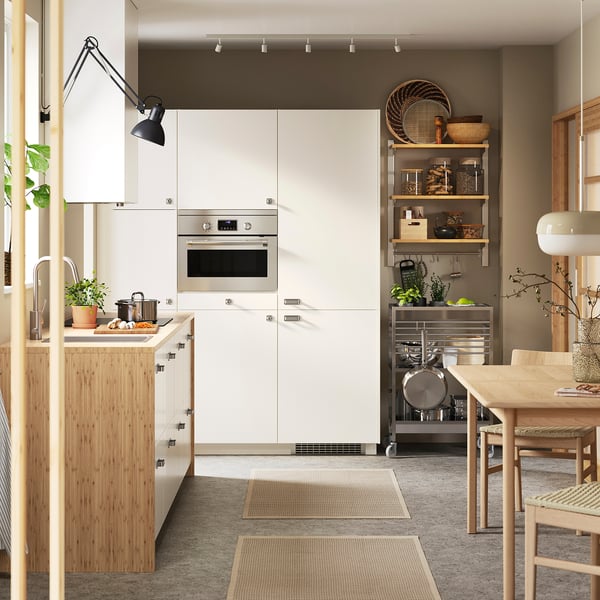 Modern kitchen with white cabinets and silver oven. Wooden dining table and chairs, potted plants, and hanging light.