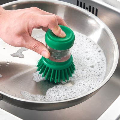 A hand holds a green brush dipping it into a stainless steel sink with soapy water, demonstrating the brushs ability to dispense soap.