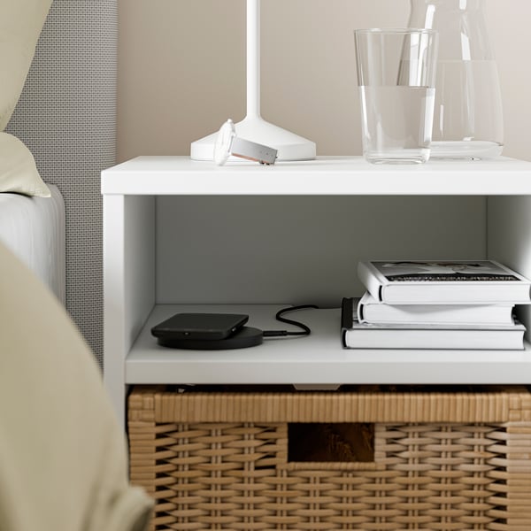 Displays a compact white VIHALS nightstand with a wicker basket and a charging phone, near a bed.