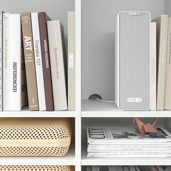 White bookshelf with seasons books, art books, speaker, basket, and origami ornament.