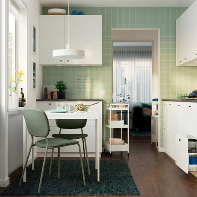 Modern kitchen with green tiles, white cabinets, wooden floor, and small VIHALS table set for two.