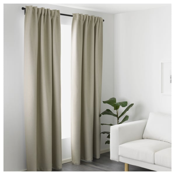 Room darkening curtains, cosy atmosphere. Beige fabric offers privacy, light reduction. White armchair, plant, minimalist decor.