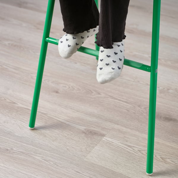 Persons feet with heart-patterned socks rest on green junior chair legs, showcasing its footrest design for children.