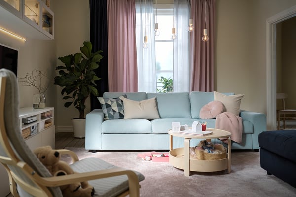 A light blue VIMLE sofa with geometric pillows in a living room. It has a large window with pink curtains and a wooden coffee table.