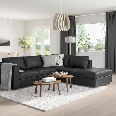 Modern living room with l-shaped VIMLE black leather sofa, wooden coffee tables, potted plants, and hanging lamps.