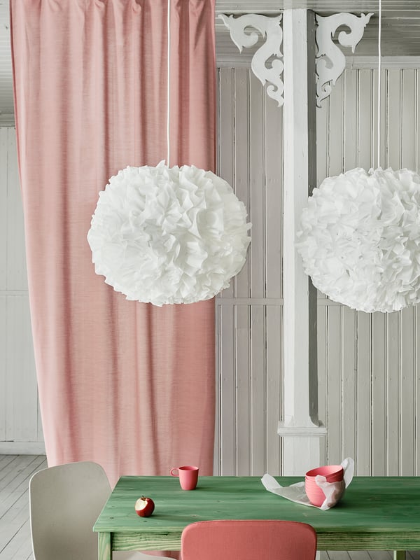 Two white spherical pendant lamps hang above a green table with a red mug and apple. Pink curtains on the left.