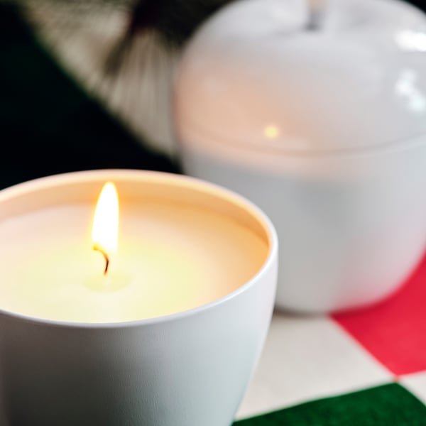A lit candle in a white cup, emitting a warm, inviting glow. Another unlit candle and a balloon partially visible nearby.