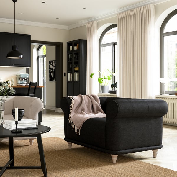 Cosy living room with VISKAFORS black armchair, beige blanket, coffee table, and bookshelf. Large windows with curtains.
