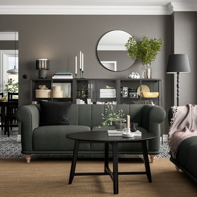 Modern living room with dark green VISKAFORS sofa, black coffee table, and grey walls. Round mirror, shelving with decor, and floor lamp.