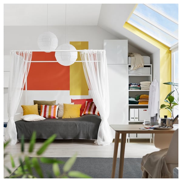 Modern grey bed framed by white curtains, styled with colourful pillows. Bright artwork, organised shelves, and wooden desk with potted plant.