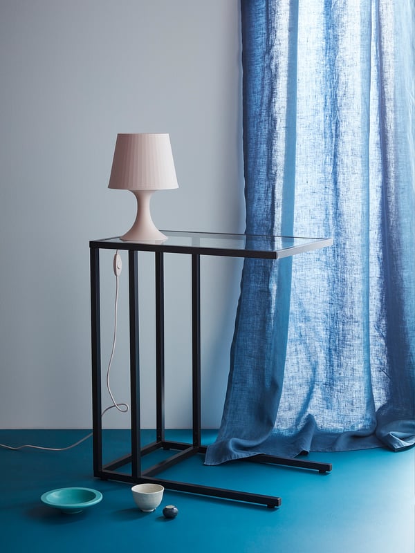 Simple table lamp on glass-top table; light blue curtain, teal floor.