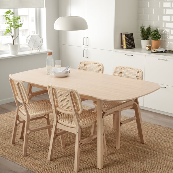 Bamboo dining set with smooth table, angled legs, & woven chairs on jute rug, white kitchen, large window.