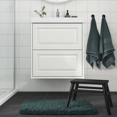 Minimalistlikus vannitoas on ALMTJÄRNi sinakasroheline vannimatt, rippuvad rätikud, valge kapp ja must taburet.