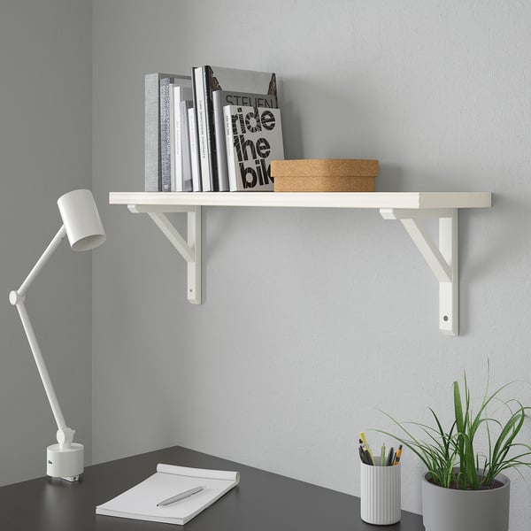 Valge riiul raamatute ja karbiga, kirjutuslaud märkmikuga, lamp, pastakad, taim.