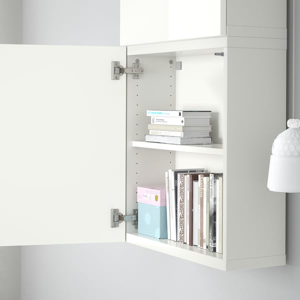 Avatud kahe riiuliga valge kapp, mis sisaldab värvilisi raamatuid ja sinist kasti. Kapp on seinale kinnitatav, osa IKEA BESTÅ hoiusüsteemist.